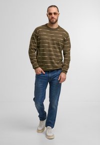 Olivengrünes Langarmshirt mit horizontalen beigen Streifen, kombiniert mit blauen Jeans und beigen Sneakers mit weißen Akzenten.