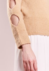 Beige gestrickter Pullover mit Rippbündchen, Ellbogen-Ausschnitten und einer weichen Textur. Kombiniert mit einem hellen Kleidungsstück.