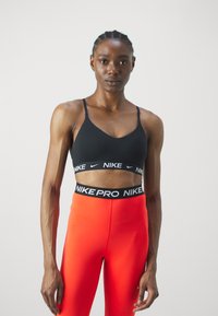 Crni sportski grudnjak s tankim naramenicama i bijelim "NIKE" logotipom na traku, u kombinaciji s visokim narančastim tajicama s crnim pojasom.