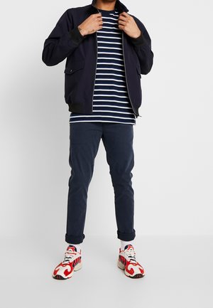 Marinblå bomberjacka med ribbade muddar, vit och marinblå randig t-shirt, mörkblå byxor och flerfärgade träningsskor med röda accenter.