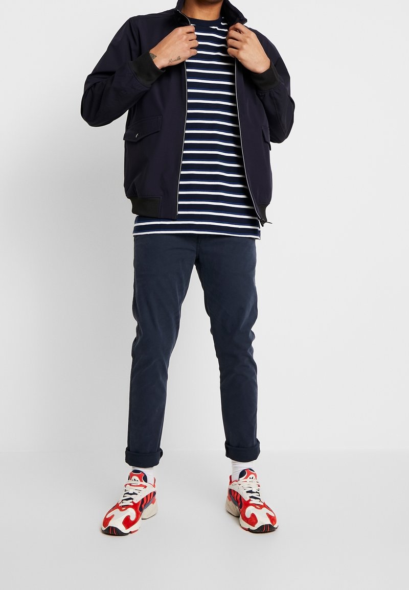 Marineblå bomberjakke med ribbede manchetter, en hvid og marineblå stribet t-shirt, mørkeblå bukser og multikolor sportssko med røde detaljer.