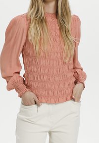 Persikofärgad blus med smockad textur, hög halsringning och puffärmar. Återfinns med krämfärgade byxor, händerna i fickorna.