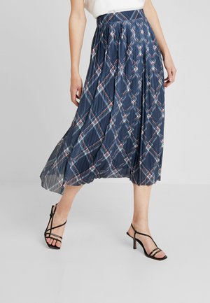 Femme portant une jupe midi plissée à carreaux bleus, associée à des sandales à talons noires à lanières, debout devant un fond gris uni.