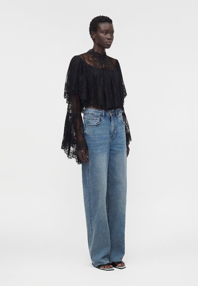 Γυναίκα που φοράει μακρυμάνικο μαύρο δαντελένιο μπλουζάκι με φαρδιά μανίκια και ψηλό λαιμό, συνδυασμένο με ανοιχτό μπλε παντελόνι φαρδιάς γραμμής και σανδάλια με ανοιχτά δάχτυλα.