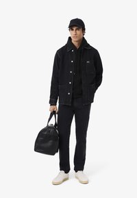 Joven con gorra negra, chaqueta de mezclilla, sudadera con capucha y pantalones, sostiene una bolsa de deporte negra, vistiendo zapatillas blancas, de pie contra un fondo blanco.