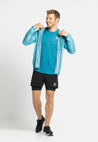 Lichtblauwe, lichte jas met capuchon over een teal-gepatrooneerd shirt en zwarte shorts. Zwarte schoenen met grijze accenten.