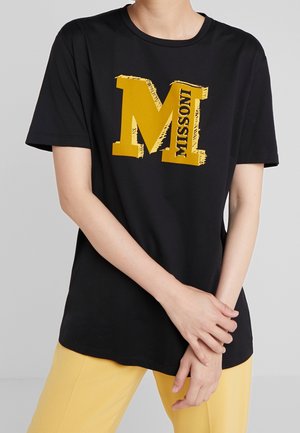 Person trägt ein schwarzes T-Shirt mit einem großen gelben Buchstaben M und dem Wort "MISSONI" auf der Vorderseite, kombiniert mit gelben Hosen.