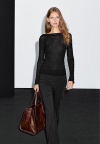 Black textured long-sleeve top paired with black fitted pants. A model holds a large, dark brown leather tote bag. Simple, modern design.