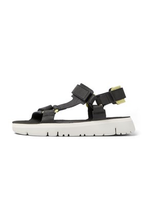 Black sandal with adjustable straps, white thick sole, textured upper material, and yellow accents. Features minimalistic and open design.