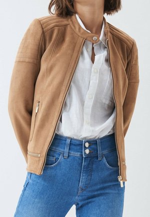 Frau trägt eine beige Reißverschlussjacke über einem weißen Hemd mit Knöpfen und hoch taillierte blaue Jeans mit doppelten Knöpfen.