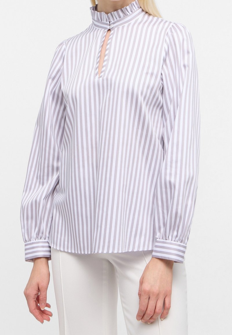 Blouse rayée blanc et lavande avec un col montant, une ouverture en forme de goutte d'eau à l'avant, et des manches longues légèrement bouffantes. Le tissu est lisse et léger.