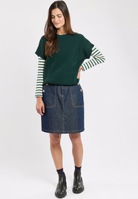 Donkergroen gebreid trui met korte mouwen over een gestreepte langemouwen shirt, gecombineerd met een donkere spijkerrok en zwarte enkellaarsjes.