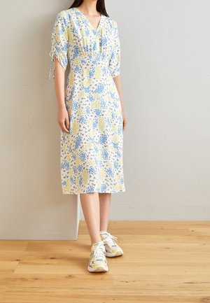 Femme portant une robe midi blanche avec des motifs floraux bleus et jaunes, ainsi que des baskets blanches et jaunes, debout sur un sol en bois.