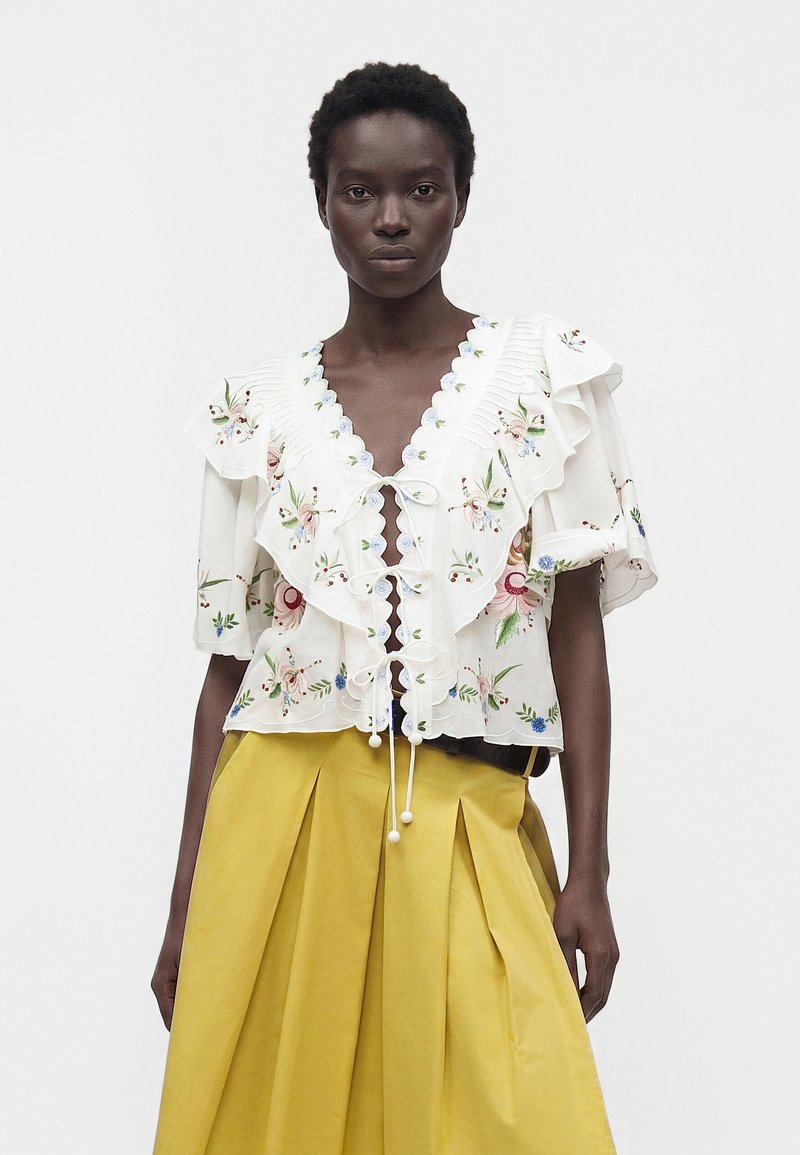 Femme portant un chemisier blanc brodé de fleurs avec des volants et des liens en dentelle, associé à une jupe plissée taille haute jaune moutarde.