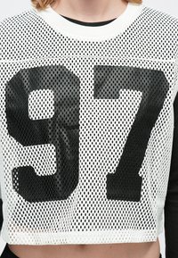 Witte mesh crop top met een grote zwarte "97" graphic. De halslijn is rond en de stof heeft een ademende geperforeerde textuur.