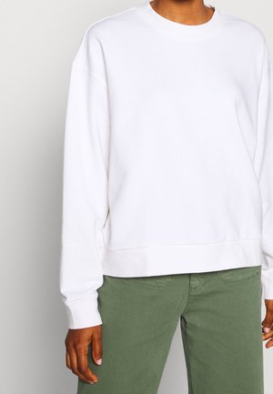Personne portant un sweat-shirt blanc uni à manches longues et un pantalon vert olive, visible du cou à la mi-cuisse, debout devant un fond neutre.