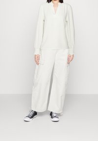 Helle cremefarbene Bluse mit V-Ausschnitt und gerafften Ärmeln, kombiniert mit weiten beigen Cargo-Hosen und schwarzen Sneakers.