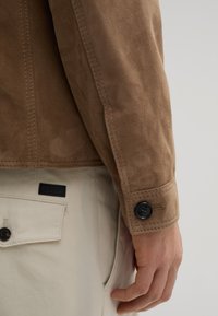 Braune Wildlederjacke mit einem Knopfverschluss am Ärmelsaum, sichtbaren Nähten und einer glatten Textur. Beigefarbene Hose mit einem Etikett an der Tasche.