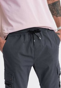 Muž v šedých cargo kalhotech s předními kapsami, černým stahovacím páskem a světle růžovou košilí, se rukama v kapsách, viditelnými tattoy na pažích a bílými hodinkami.