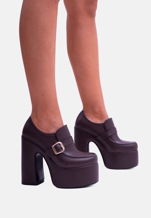 Scarpe platform in finta pelle marrone scuro con punta arrotondata, tacco chunky e dettaglio con fibbia. Texture liscia e design moderno.