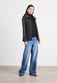 Een vrouw met lang, donker haar draagt een zwarte gewatteerde jas en een coltrui, gecombineerd met een wijdvallende blauwe jeans, staand voor een neutrale achtergrond.
