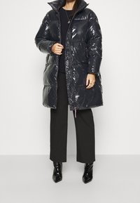 Svart glänsande puffjacka med hög krage, dragkedja och sidofickor. Bärs över svarta byxor och kombineras med blanka svarta stövlar.