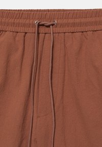 Pantalon en tissu marron avec taille élastique et cordon marron noué à travers deux œillets métalliques au centre devant.