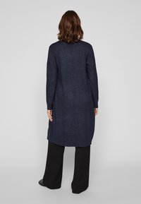 Μακρύ ναυτικό καζάκα από υφασμένο πλεκτό ύφασμα, με ίσια γραμμή και πλάγιες σχισμές, φορεμένη πάνω από μαύρες φαρδιές παντελόνες.