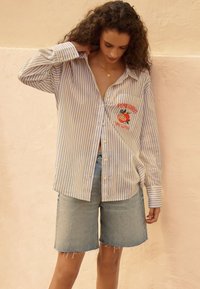 Striped button-up shirt with blue and white stripes, featuring a pocket with red embroidered text and a graphic. Paired with denim shorts.