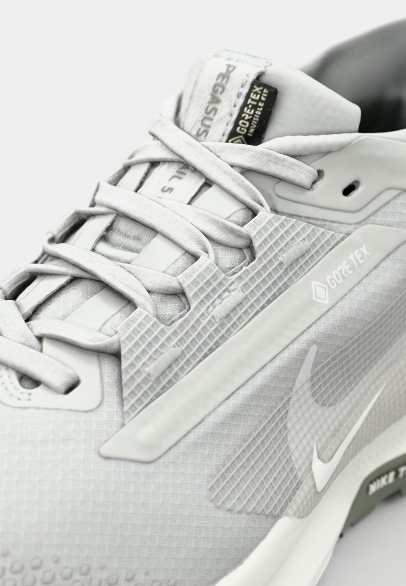 Zapatilla de running color gris claro con parte superior texturizada, que presenta la etiqueta GORE-TEX, suela de goma y cordones en tono. Logo de Nike en el costado.
