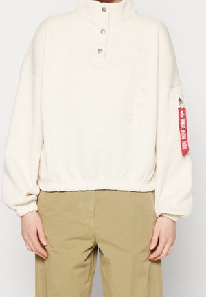 Pull écru avec col montant, boutons-pression, ourlet et poignets élastiques, texte embossé "Alpha Industries" et étiquette rouge "Remove Before Flight" sur la poche de la manche.