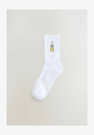 Weiße Baumwollsocken mit geripptem Muster, die auf der Seite eine bestickte gelbe Fanta-Flasche zeigen.