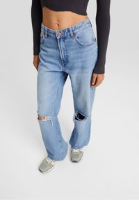 Donna che indossa jeans strappati di un blu chiaro, sneakers grigie e un top cropped nero a maniche lunghe su uno sfondo semplice.