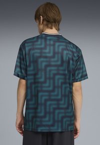 Persona con capelli castani corti che indossa una maglietta a maniche corte con motivo a zigzag nero e verde bluastro, vista da dietro su uno sfondo grigio.