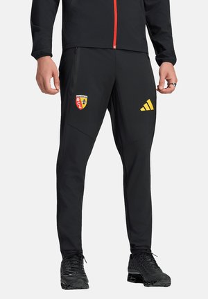 Uomo che indossa pantaloni sportivi neri con il logo del Racing Club de Lens sulla coscia sinistra e il logo giallo Adidas sulla coscia destra, abbinati a sneaker nere.