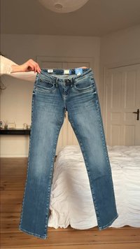 Osoba drží modré rifle za pás v spálni s drevenou podlahou a bielym prehozom na posteli.