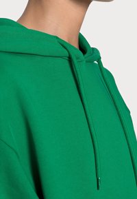 Sudadera verde con cordón, confeccionada en tejido suave. Presenta un ajuste holgado y puños acanalados, mostrando una textura lisa.