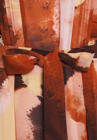 Vaatteena oleva kietaisumekko, jossa on teksturoitu kangas monivärisellä abstraktilla kuviolla lämpimissä sävyissä. Mekko on varustettu ruskealla vyötärösoljella.