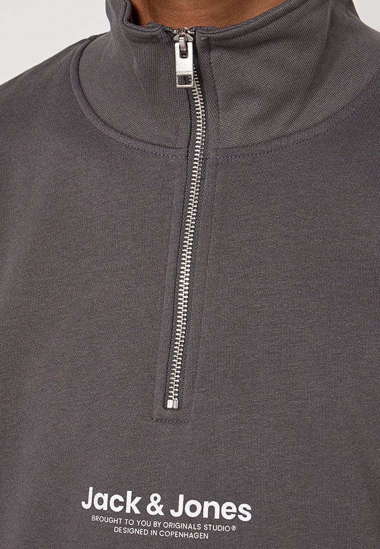Sudadera gris con cremallera y cuello alto, cremallera plateada, material de algodón y texto de logo blanco en la parte inferior. Diseño simple y moderno.
