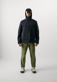 Veste noire imperméable avec capuche, associée à un pantalon vert. Des lunettes de soleil réfléchissantes et des chaussures de randonnée beiges complètent la tenue.