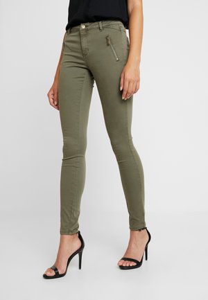 Vrouw draagt slank gesneden olijfgroene broek met voorzakken met rits en zwarte sandalen met hoge hak, staand tegen een effen achtergrond.