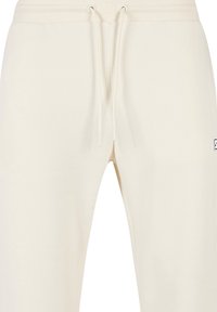 Pantalones de chándal color crema hechos de una tela suave, con una cinturilla elástica con cordones y un pequeño parche de logo en el costado.