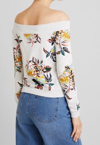 Felpa floreale a spalle scoperte di colore grigio chiaro con motivi di fiori colorati. Tessuto morbido, colletto e polsini a coste, abbinata a jeans blu.