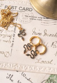 Guld slange-inspirerede øreringe og matchende halskæde, med indviklede design med blå accenter, hvilende på vintage cremefarvet papir.