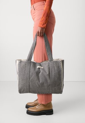 Person, der holder en stor sort-hvid mønstret tote-taske med tykke stropper, iført ferskenfarvede bukser, orange top og lysebrune snørestøvler med sorte såler.