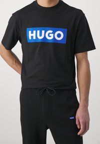Schwarzes Baumwoll-T-Shirt mit einem großen blauen rechteckigen grafischen Element, auf dem das weiße Wort "HUGO" zentriert ist. Kombiniert mit schwarzen Jogginghosen mit Kordelzug.