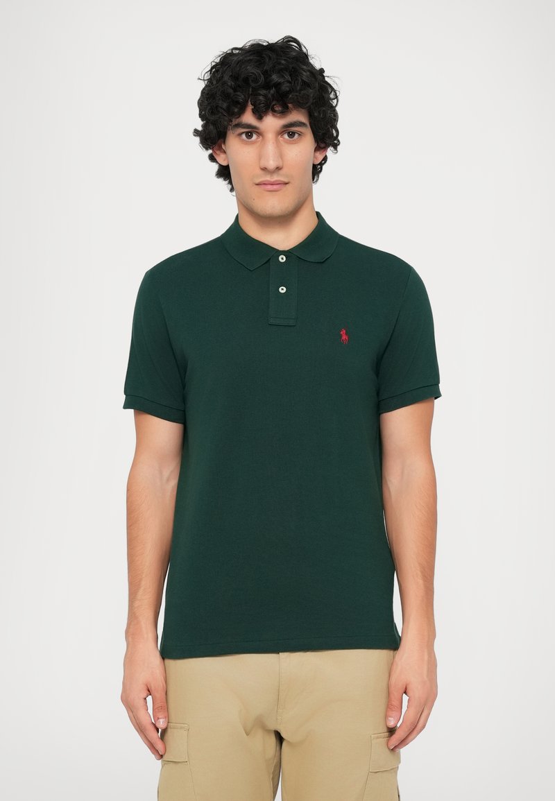 Mörkgrön polo t-shirt med krage, korta ärmar, två knappar och en röd broderad logga på vänster bröst. Mjuk bomullstyg.