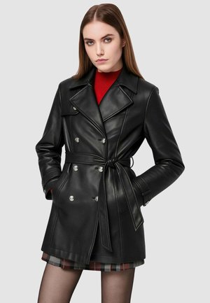 Cappotto doppiopetto in ecopelle nera con rever a cuneo, vita con cintura e bottoni argento. Indossato sopra una maglia rossa e una gonna a fantasia.