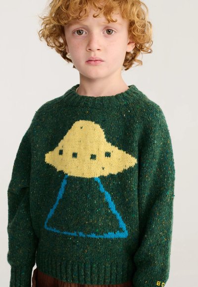 Enfant aux cheveux roux bouclés portant un pull vert avec un grand motif jaune d'OVNI avec des rayons bleus sur le devant.