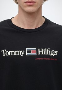 Νέος/νέα που φοράει μαύρο T-shirt με λευκό και κόκκινο λογότυπο Tommy Hilfiger και το κείμενο "Authentic Originals since 1985" στο μπροστινό μέρος.